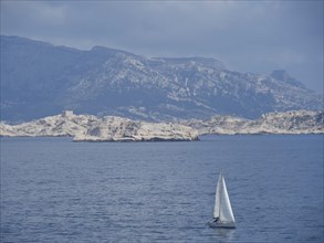 Sailboat sailing on a calm sea in front of a rocky mountain landscape and cloudy sky, Marseille on
