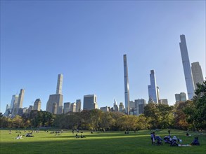 A sunny day with people on the grass in central park, surrounded by tall skyscrapers, the skyline