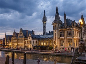 Gothic buildings with illuminated windows and people walking along a canal under a cloudy sky,