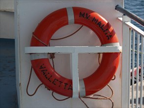 A bright orange lifebuoy with lettering on board a boat, the island of Gozo with historic houses,