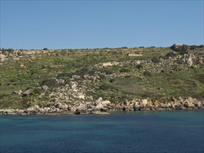 A rocky coastline with green hills, clear waters and sunny skies, the island of Gozo with historic