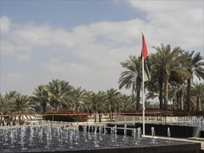 A fountain surrounded by palm trees under a slightly cloudy sky with a waving flag in the