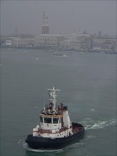 A tugboat on calm water, background shows foggy historic buildings of the city, the lagoon city of