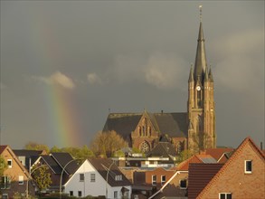 A gothic church towers over the city view in a cloudy sky with a rainbow in the background, rainbow