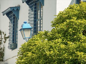 White building with blue shutters and bars, next to it a blue street lamp and green bushes,
