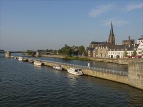River view with neighbouring boats, historic buildings and a church, a bridge in the distance,