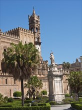 Historic cathedral with a high tower and adjoining statue, surrounded by palm trees under a clear
