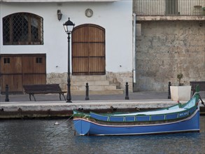 A boat on the water in front of a building with doors and windows, lantern on the shore, Valetta,