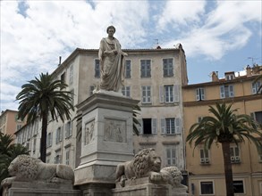 City monument with statue and lion surrounded by historic buildings and palm trees, Corsica,