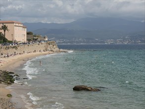 Beach along the clear blue sea with buildings, palm trees and mountains in the background under a