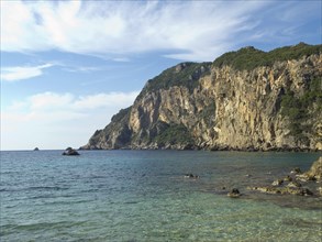 Rocky beach with clear blue water and steep cliffs against a partly cloudy sky, beach on the island