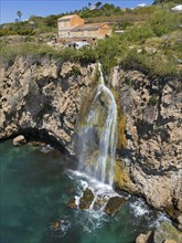 A picturesque waterfall falls from a cliff into turquoise water with houses in the background, a