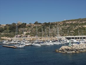 Marina with many sailing boats and adjacent hills under a bright blue sky, the island of Gozo with