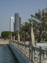 An urban promenade with palm trees along the water and tall buildings in the background, Abu Dhabi,