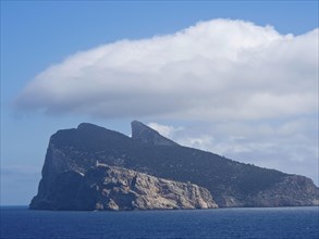 A rocky island rises against a clear blue sky with gentle clouds over the calm sea, palma de