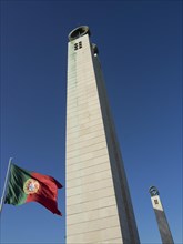 A tall tower with a Portuguese flag in the foreground against a blue sky, Lisbon on the Tagus river