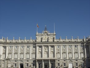 The picture shows the symmetrical facade of a palace with a flag under a clear blue sky, Madrid,