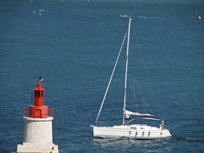 A sailing boat sails in the blue sea next to a red and white lighthouse, la seyne sur mer, france
