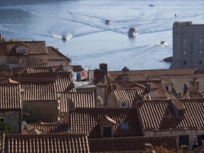 View over red tiled roofs to the sea with boats and some buildings on the coast, the old town of