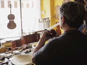 From behind, a luthier works intently on a cello in a well-organized workshop with natural light