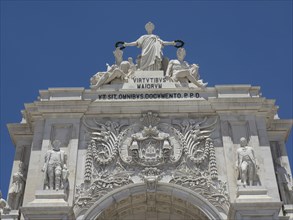 Close-up of the upper part of a triumphal arch with detailed statues and inscription against a blue
