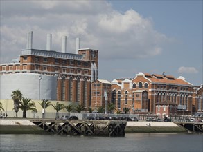 Industrial buildings with palm trees in front of a body of water under a clear sky, Lisbon,