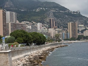 Coastline with promenade, skyscrapers and mountains behind, surrounded by sea and cloudy sky,