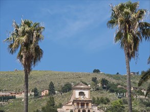 Two tall palm trees frame a church in front of a green hilly landscape under a blue sky, palermo in