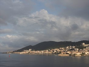 City by the sea with many buildings, behind it a hilly landscape under a cloudy sky, Corsica,