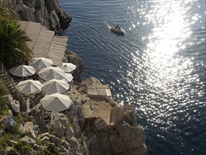 View of the sea from the rock with white parasols and a boat on the water, reflecting sun rays, the