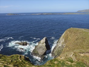 Impressive cliff landscape with rough waves crashing against rocks, under a blue sky, green meadows