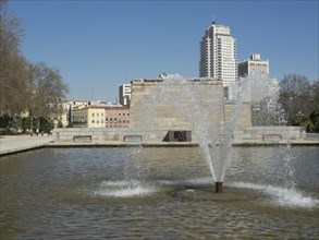 Fountain with water jets in front of historic buildings in a park under a blue sky, Madrid, Spain,