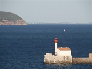 White and red lighthouse on a wall in the sea with a distant island and blue sky, la seyne sur mer,