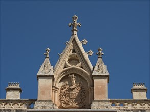 Detailed pediment with ornate coat of arms and elegantly designed spires under a clear blue sky,