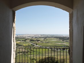 Panoramic view of a wide landscape and a town, seen through a stone archway, the town of mdina on