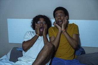Scared gay couple with hands on face and open mouth watching horror movie at home