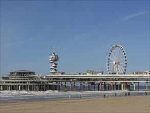 Pier with Ferris wheel and tower on the beach, people walking along the beach, parasols and a pier