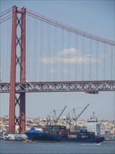 Red suspension bridge over the water, below container harbour with large cargo ships, Lisbon,