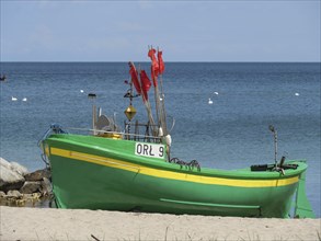 A green boat with red flags stands on the beach, propped up on the sand, in front of a calm sea,