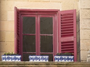 Red window with shutters and flower pots on a stone wall, the town of mdina on the island of malta