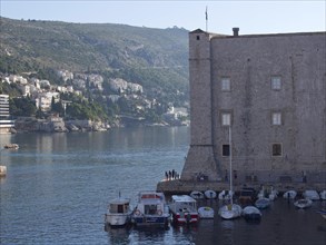 Stone building at the edge of a harbour with boats in the water, city and mountains in the