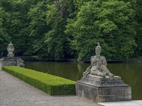 Stone statue in a park next to a lake, surrounded by dense green trees, historic castle with a