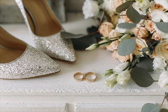 Elegant glitter shoes and wedding rings placed near a wedding bouquet of white flowers and peach