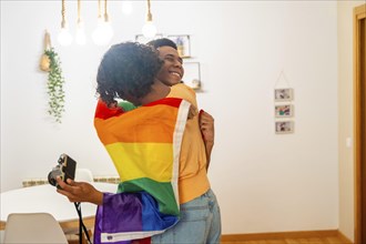 Gay latin young couple embracing wrapped in LGBT flag while taking photos in the apartment