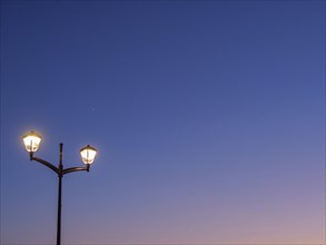 A street lamp shines against a dark blue sky at dusk, sunset in many colours on a beach with a calm
