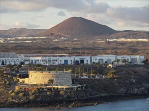 A castle and modern buildings on the coast, mountains and a volcano in the background, Lanzarote,
