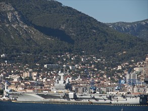 A large warship in the harbour with a town and hills in the background under a clear sky, la seyne