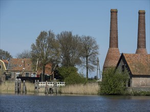 A picturesque river with old buildings and chimneys in the background, surrounded by trees,