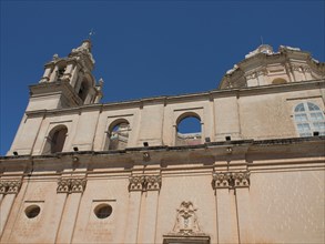 Baroque church facade with decorative elements and two towers against a blue sky, the town of mdina