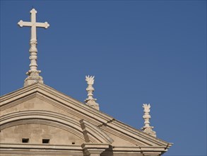 Top of a church with a cross and classical style elements in front of a cloudless blue sky, the old